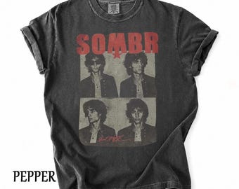 May include: A charcoal gray short-sleeve t-shirt featuring a red graphic print with the text SOMBR at the top. The design includes four black and white photographic portraits of a person with wavy hair, arranged in a grid, with a red signature below.
