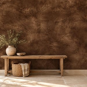 May include: A long wooden bench sits against a dark brown textured wall. On the bench, there is a light-colored ceramic vase containing green olive branches, a small bowl, and a woven basket containing a folded white and brown striped cloth.