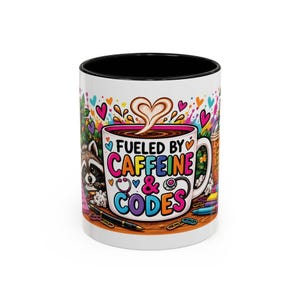 May include: A white ceramic mug with a black interior and rim, featuring a colourful graphic of a coffee cup with heart-shaped foam. The text reads FUELED BY CAFFEINE & CODES, surrounded by illustrations of a raccoon, pens, and paper clips.