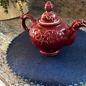 May include: A deep red ceramic teapot featuring an embossed floral pattern on the body and lid. The teapot has a curved handle, a spout, and a matching lid with a pointed finial. The surface of the teapot is glossy and reflects light.