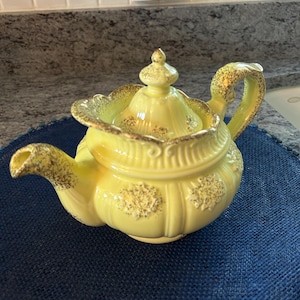 Puede incluir: Una tetera de cerámica de color amarillo pálido con tapa extraíble, pomo, asa curva y pico. La tetera está decorada con patrones florales en relieve y motas de color dorado a lo largo de los bordes, el asa, el pico y el borde de la tapa.
