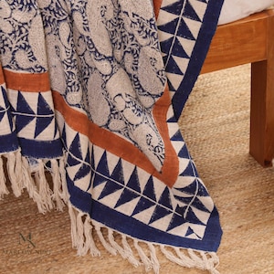 May include: A woven textile throw featuring a cream-colored base with a dark blue paisley pattern, accented by a solid burnt orange stripe and a dark blue border with a repeating triangle motif. The edges are finished with cream-colored fringe.