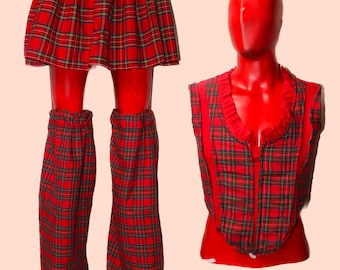 Könnte beinhalten: Ein rotes Karo-Outfit bestehend aus einem Faltenrock, einer ärmellosen Weste mit gerüschtem Ausschnitt und einem Paar passender Stulpen. Der Stoff weist durchgehend ein rotes, grünes und gelbes Schottenkaro-Muster auf.