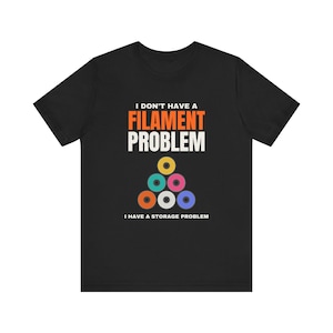 May include: A black short-sleeve t-shirt featuring the text I DON'T HAVE A FILAMENT PROBLEM I HAVE A STORAGE PROBLEM in white and orange, with a graphic of six colorful 3D printer filament spools arranged in a pyramid shape.