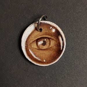 May include: A circular wooden pendant featuring a hand-painted brown eye design with a glossy finish. The pendant has a silver-toned metal jump ring attached at the top for hanging, with visible wood grain texture and white highlights.