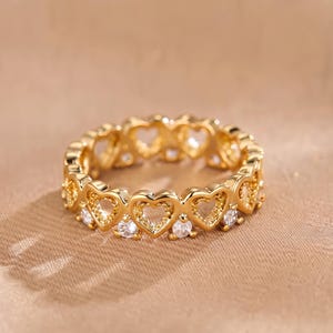Puede incluir: Un anillo de tono dorado que presenta una banda continua de recortes en forma de corazón. Pequeñas piedras transparentes de corte redondo están engastadas entre cada motivo de corazón, creando un patrón repetitivo de corazones metálicos.
