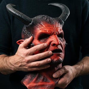 May include: A red and black devil mask featuring prominent curved horns, textured skin details, deep-set eye sockets, and a defined facial structure. The mask displays a weathered appearance with dark shading around the features and pointed ears.
