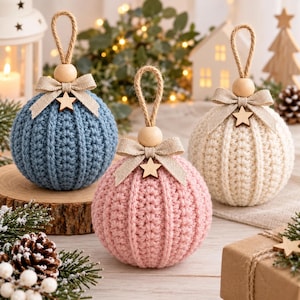 Puede incluir: Tres adornos esféricos con un patrón de tejido de ganchillo texturizado en azul, rosa y crema. Cada adorno está rematado con una cuenta de madera natural, un lazo de tela beige y una pequeña estrella de madera, con un lazo de yute.
