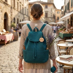 May include: A teal crochet backpack with a flap closure, drawstring, and two side pockets. The bag features a round wooden tag with the text Etsy and a small Etsy seller pin on the jacket. A bundle of dried lavender is tucked into one side pocket.
