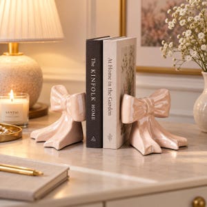 May include: A pair of glossy, light pink ceramic bookends shaped like decorative bows. The bookends are positioned on either side of two books, one titled The Kinfolk Home and the other titled At Home in the Garden, holding them upright.