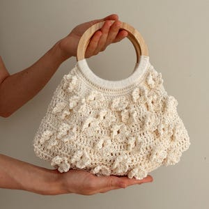 May include: A cream-coloured crochet handbag featuring a textured floral pattern across the body and a round, light-toned wooden handle. The bag is constructed with intricate yarn stitching and has a structured, circular opening at the top.