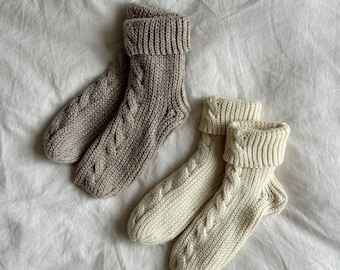 May include: A pair of knitted socks in a light beige color and a pair of knitted socks in an off-white color. Both pairs feature a vertical cable knit pattern running down the center and a thick, folded ribbed cuff at the top of each sock.
