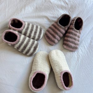 May include: Three pairs of fuzzy slippers. One pair features white and brown horizontal stripes with pink trim. Another pair has brown and pink horizontal stripes. The third pair is solid cream-colored with pink trim around the ankle openings.