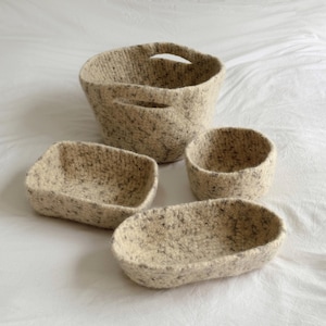 Op de afbeelding: Een set van vier beige en grijs gespikkelde vilten bakjes, inclusief een grote ronde mand met twee geïntegreerde handvatten, een kleine ronde kom, een rechthoekig dienblad en een ovaal dienblad, allemaal met een getextureerd oppervlak.