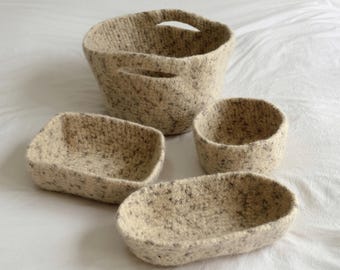 May include: A set of four beige and grey speckled felted containers, including one large round basket with two integrated handles, one small round bowl, one rectangular tray, and one oval tray, all featuring a textured, soft-looking surface.