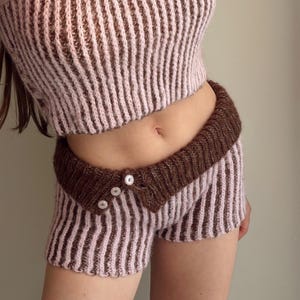 Könnte beinhalten: Ein zweiteiliges Strickset bestehend aus einem ärmellosen bauchfreien Oberteil und einer Shorts. Beide Teile haben ein vertikales Rippenmuster aus abwechselnd hellrosa und dunkelbraunem Garn. Die Shorts hat einen breiten, dunkelbraunen Umschlagbund mit drei Knöpfen.