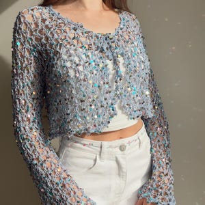 May include: A light blue open-front crochet cardigan featuring long sleeves and a loose, net-like knit pattern. The garment is embellished with numerous small, iridescent, multi-colored sequins that reflect light throughout the entire piece.
