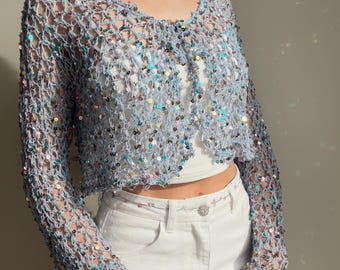 May include: A light blue open-front crochet cardigan featuring long sleeves and a loose, net-like knit pattern. The garment is embellished with numerous small, iridescent, multi-colored sequins that reflect light throughout the entire piece.