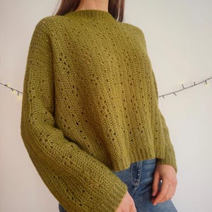 May include: A long-sleeved olive green crochet sweater featuring a ribbed crew neckline and cuffs. The garment is crafted with a repeating vertical pattern of openwork stitches and solid crochet columns, creating a textured, semi-sheer appearance.