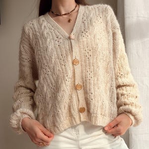 Peut inclure: Un cardigan couleur crème à col en V présentant un motif torsadé texturé, des manches longues avec des poignets côtelés et quatre boutons ronds translucides sur le devant. Le tricot possède une structure légèrement ouverte et aérée.