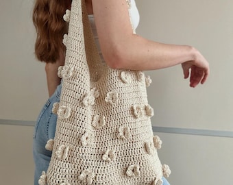 May include: A cream-colored crochet tote bag featuring a textured pattern of small, raised floral appliques distributed across the front and along the shoulder strap. The bag is constructed with a visible knit stitch and has a wide, open top.