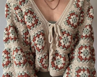 May include: A long-sleeved crochet cardigan constructed from cream, sage green, and terracotta granny squares. The garment features a deep V-neckline, a front tie closure, and a loose, relaxed fit with wide sleeves, showcasing intricate yarn work.
