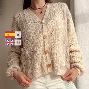 May include: A cream-colored V-neck cardigan featuring a cable knit pattern, long sleeves with ribbed cuffs, and four round buttons down the front. The garment includes text overlays reading ES and EN next to small flag icons.
