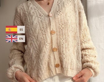 May include: A cream-colored V-neck cardigan featuring a cable knit pattern, long sleeves with ribbed cuffs, and four round buttons down the front. The garment includes text overlays reading ES and EN next to small flag icons.