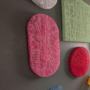 Op de afbeelding: Een verzameling wandkunstwerken van gespannen draad in diverse vormen en kleuren, waaronder een grote roze ovaal, een groen vierkant, een rode cirkel, een gele ovaal, een witte cirkel, een blauw vierkant en een kleine gele bol op een donkere muur.