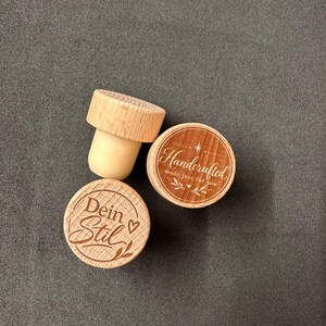 Pode incluir: Dois rolhas de garrafa redondas de madeira com bases cónicas de cor clara. Uma rolha apresenta o texto gravado "Handcrafted made just for you" com um design de estrela e folhas. A outra rolha exibe o texto gravado "Dein Stil" com um coração.