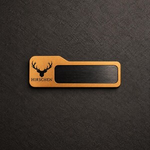 May include: A rectangular name badge featuring a gold-toned finish with a black deer head silhouette and the text HIRSCHEN printed below it. The badge includes a central horizontal black rectangular insert with a brushed metal texture.