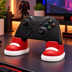 May include: A black video game controller resting in a red and white sneaker-shaped stand. The controller features two analog sticks, a directional pad, and four colored action buttons labeled A, B, X, and Y. The stand supports the controller.