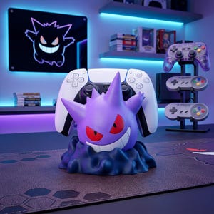 May include: A purple Gengar character figure serving as a controller stand, featuring red eyes and a wide grin, supporting a white and black wireless game controller. The figure is mounted on a dark, textured base resembling a cloud of smoke.