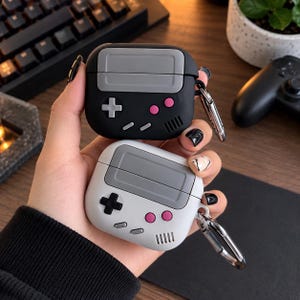 Può includere: Due custodie protettive in silicone per auricolari wireless, progettate per assomigliare a console di gioco portatili. Una custodia è nera con un'area dello schermo grigia, un pad direzionale grigio chiaro, due pulsanti rosa e due piccoli tasti grigi.