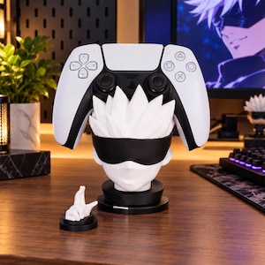 Puede incluir: Un soporte para mando de videojuegos en blanco y negro con forma de cabeza de personaje de anime, con pelo puntiagudo y una venda negra. El soporte tiene una base circular y se acompaña de una pequeña figura blanca de un gesto manual.
