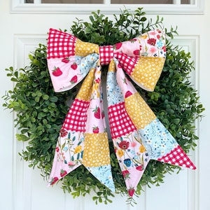 May include: A green boxwood wreath adorned with a large, multi-patterned fabric bow. The bow features a patchwork design of red and white gingham, pink fabric with strawberries, yellow floral prints, and light blue wildflower patterns.