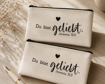 Könnte beinhalten: Zwei rechteckige Federmäppchen aus beigem Canvas mit schwarzen Reißverschlüssen. Jedes Mäppchen ist mit einem schwarzen Herzsymbol und dem aufgedruckten Text Du bist geliebt. Jeremia 31.3 in einer Kombination aus Serif- und Schreibschrift.