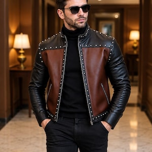 Puede incluir: Una chaqueta de dos tonos con mangas de cuero negro y un panel de pecho de cuero marrón, acentuada con tachuelas de metal plateado a lo largo de las costuras y el cuello. La chaqueta se lleva sobre un suéter de cuello alto negro y pantalones negros.