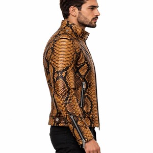 K&ouml;nnte beinhalten: Eine Jacke mit braun-schwarzem Schlangenledermuster, Stehkragen, silberfarbenen Metallrei&szlig;verschl&uuml;ssen an der Vorderseite und an den &Auml;rmelb&uuml;ndchen sowie Druckknopfdetails an Schultern und Saum, getragen &uuml;ber einer schwarzen Hose.