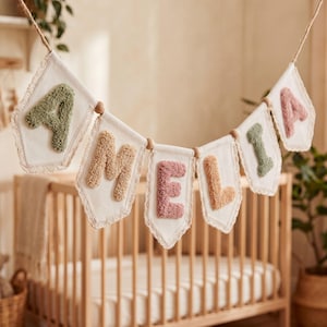 May include: A decorative banner featuring the name AMELIA spelled out in individual fabric pennants. Each pennant is off-white with lace trim and displays a single tufted letter in muted shades of sage green, tan, and dusty rose.