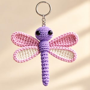 May include: A crochet dragonfly keychain featuring a purple head and body with large black eyes. The four wings are outlined in pink yarn with cream-colored centers. A silver metal split ring is attached to the top of the dragonfly head.