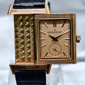 May include: A rectangular rose gold wristwatch with a textured gold dial, black hands, and a sub-dial. The watch face features the text Jaeger-LeCoultre and Swiss Made. It has a black alligator-patterned leather strap and a metallic side panel.