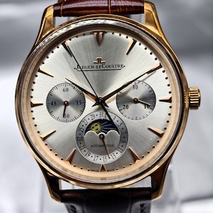 Può includere: Un orologio da polso Jaeger-LeCoultre color oro con quadrante argentato a raggiera, indici in oro rosa e cinturino in pelle marrone con motivo alligatore. L'orologio presenta due sotto-quadranti, una fase lunare e la scritta Jaeger-LeCoultre Automatique.