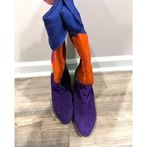 May include: A pair of knee-high boots featuring a multi-colored suede design. The boots have a deep purple toe and lower shaft, transitioning into bright orange, green, and blue panels along the upper section with a slouchy fit.