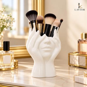 Peut inclure: Un porte-pinceaux de maquillage en céramique blanche en forme de mains entourant un visage. Le support contient plusieurs pinceaux de maquillage avec des manches noirs et des poils bruns et blancs. Le texte LAVIOR est visible en haut à droite.