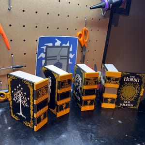 May include: A set of four 3D-printed book-shaped storage containers featuring black and yellow designs. The spines are labeled The Return of the King, The Two Towers, The Fellowship of the Ring, and The Hobbit, with matching cover art.