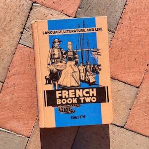 May include: A vintage French textbook titled "French Book Two" with the subtitle "Language, Literature, and Life" by Smith. The cover features a colorful illustration of people in traditional French clothing standing in front of a harbor scene.
