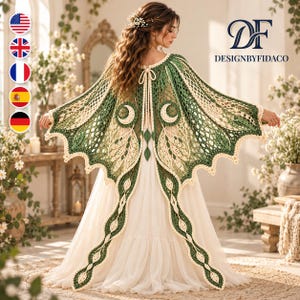 May include: A crochet cape featuring a green and cream color scheme with intricate wing-like patterns, crescent moon motifs, and a tie closure at the neck. The garment is worn over a long, plain white dress, showcasing detailed lace-like stitching.