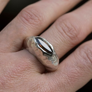 Puede incluir: Un anillo de plata con una banda texturizada y una piedra ovalada marr&oacute;n.