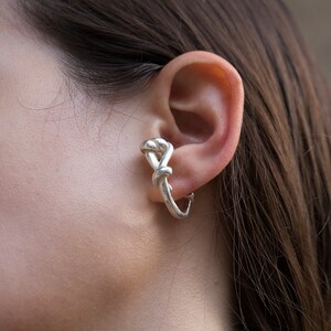 May include: Silver ear cuff with a knotted design. The cuff is worn on the upper part of the ear.